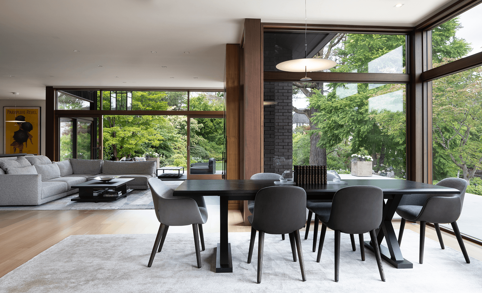 A modern dining table sits in the foreground of an open concept living area with floor-to-ceiling windows.